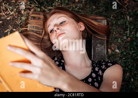 Teen, ragazza dello studente dello zenzero che si trova sulla panchina, leggendo un libro Foto Stock