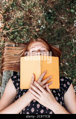 Teen, ragazza dello studente dello zenzero che si trova sulla panchina, leggendo un libro, guardando molto felice Foto Stock