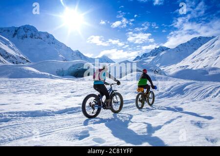 Due donne in bicicletta grasso di fronte al ghiacciaio di Skookum, Chugach National Forest, Alaska in una giornata di sole inverno, pugno pompando mentre lei corre Foto Stock