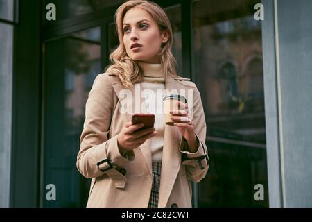 Bella ragazza bionda casual in trench elegante cappotto con caffè da andare e cellulare all'aperto Foto Stock