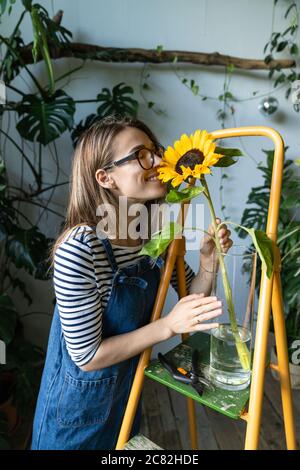 Donna fiorista circondata da piante tropicali gode di un bel girasole in vaso, in piedi vicino alla scala arancione. Foto Stock