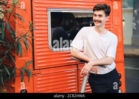 Giovane gioioso baffi uomo con la T-shirt appoggiata su e-scooter felice di guardare da parte con furgone rosso sullo sfondo della strada Foto Stock