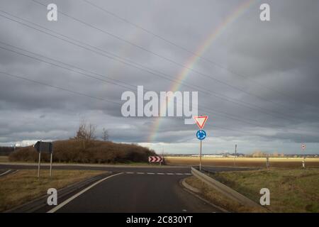 Doppio arcobaleno nel cielo nuvoloso su un campo e. una strada con rotatoria e indicazioni per il rendimento Foto Stock