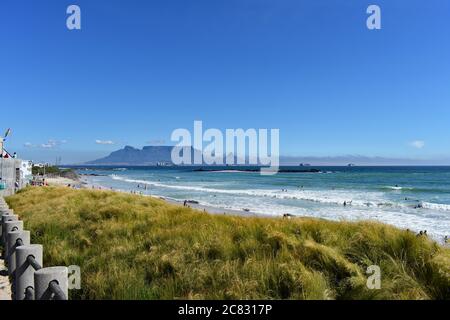 Table Mountain, Devils Peak e Lions Head visti da Bloubergstrand Beach da tutta Table Bay. L'erba verde contrasta con l'azzurro del cielo e dell'oceano Foto Stock