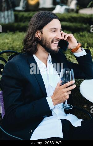 Giovane bruna sorridente portava uomo d'affari con un bicchiere di vino rosso che parlava felicemente sul cellulare nel ristorante all'aperto Foto Stock