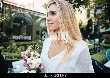 Giovane bella donna romantica in abito bianco con bouquet di fiori sensualmente guardando da parte nel giardino del ristorante Foto Stock