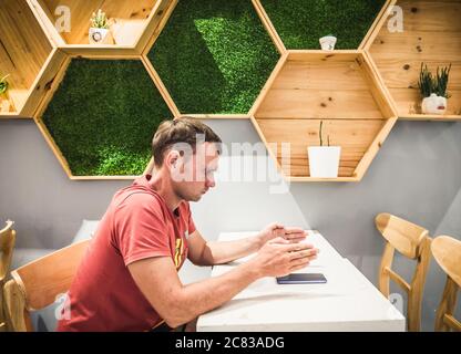 Serio bell'uomo biondo in stile casual t-shirt arancione seduta in un moderno caffè con telefono cellulare di fronte a lui sul tavolo, mostra la cornice con Foto Stock