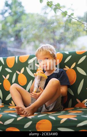 Vista frontale del ragazzo biondo felice assetato che beve limonata, tè alla frutta, tè al ghiaccio kombucha con pesca e limone, bevande all'arancia benessere da tazza di plastica Foto Stock