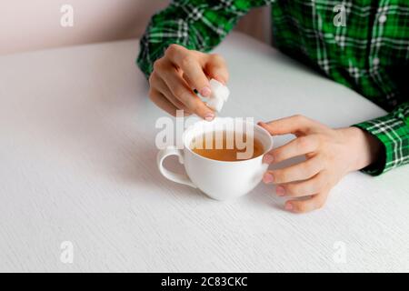 una mano di persona che tiene alcuni cubetti di zucchero e li mette in tazza di tè seduto al tavolo Foto Stock