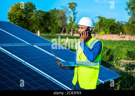 giovane tecnico maschile lavoratore che indossa giubbotti verdi e casco con laptop in mano parlando al telefono sulla manutenzione e l'installazione del solare p Foto Stock