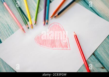 Primo piano di un disegno a cuore rosso su un bianco carta con matite colorate sul tavolo Foto Stock