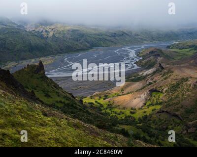 Vista dalla cima della montagna Valahnukur sul paesaggio della verde valle di Thorsmork con le rocce verdi affilate, il delta del fiume Krossa e la foresta verde. Nebbia Foto Stock