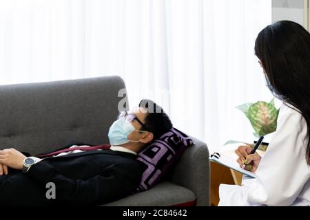 Psicoterapeuta medico femminile in visita casa paziente lavorando con depresso uomo d'affari asiatico che si trova su poltrona divano in soggiorno per consultare ab Foto Stock