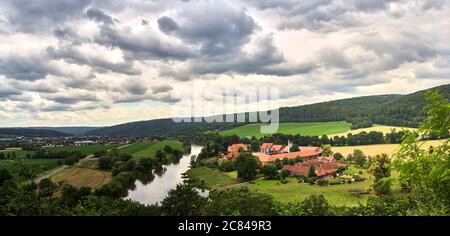 Vista da una montagna al corso del Weser con una casa padronale e un villaggio sulla riva verde con campi e prati con cielo drammatico Foto Stock