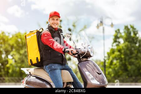 Courier Man con zaino giallo seduto sullo scooter sorridendo all'esterno Foto Stock