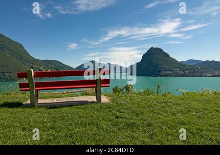 Bella vista panoramica sul Lago di Lugano (Lago di Ceresio) con panca rossa e il Monte San Salvatore sullo sfondo del Parco San Mich Foto Stock