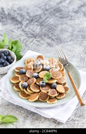 Minuscoli pancake per colazione. Frittelle di cereali con mirtilli in piastra su sfondo grigio. Cibo alla moda. Spazio di copia per testo o disegno Foto Stock