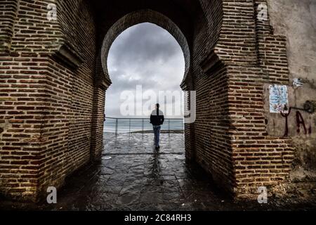 Marocco, Tangeri: Scena dalla vita quotidiana con un abitante. Giovane uomo guardato attraverso la volta costolata di una porta, sotto il cielo grigio, di fronte all'Atl Foto Stock