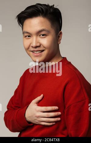 Immagine di felice bell'uomo asiatico sorridente e tenendo la mano sul petto isolato su sfondo grigio Foto Stock