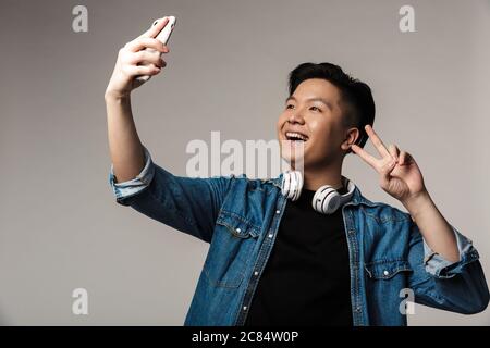 Immagine di un giovane asiatico sorridente che prende selfie su smartphone e gesturante pace cantando isolato su sfondo grigio Foto Stock