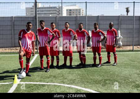 Squadra multietnica di calciatori maschili che indossano una squadra di allenamento in un campo sportivo al sole, che si pone a macchina fotografica con una palla. Foto Stock