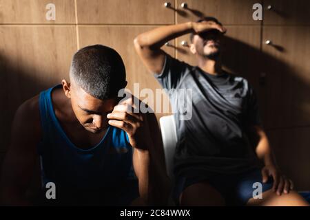 Due giocatori di calcio maschile di razza mista che indossano abiti sportivi seduti in spogliatoio durante una pausa di gioco, che si riposano tenendo la testa in disappo Foto Stock