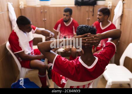 Gruppo multietnico di giocatori di calcio maschili che indossano una striscia di squadra seduto in spogliatoio durante una pausa di gioco, interagendo e parlando. Foto Stock