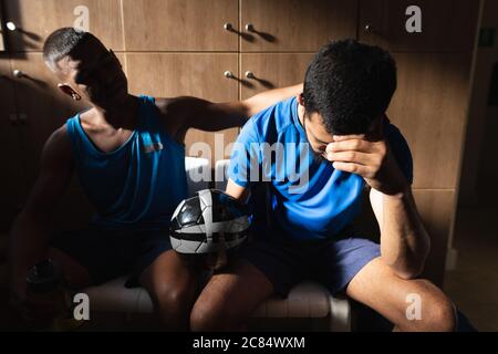 Due giocatori di calcio maschile di razza mista che indossano abiti sportivi seduti in spogliatoio durante una pausa di gioco, tenendo la palla a riposo essere deluso. Foto Stock