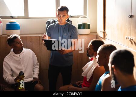 Gruppo multietnico di giocatori di calcio che indossano abiti sportivi seduti in spogliatoio durante una pausa di gioco, ascoltando il loro allenatore che istruisce ho Foto Stock