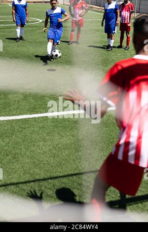 Due squadre multietniche di giocatori di calcio maschili che indossano una striscia di squadra che giocano una partita in un campo sportivo al sole, un giocatore che calcia palla portiere i Foto Stock