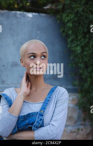 Misto razza alternativa donna con capelli biondi corti fuori e circa in città in una giornata di sole, in piedi da un muro e guardando via. Indipendente urbano w Foto Stock