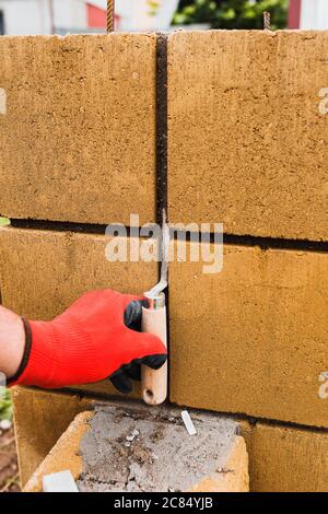 Mani di un muratore professionale con guanti mentre si lavora sulla costruzione di pareti in pietra - un cazzuola per l'applicazione di Malta in muratura e un martello per muratura Foto Stock