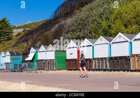 Bournemouth, Dorset UK. 21 luglio 2020. Dopo il fuoco alla spiaggia di West Cliff, Bournemouth che ha iniziato in capanna sulla spiaggia, mostrando resti di scheletro Charred resti di capanna sulla spiaggia con danni a quelli vicini e resti di scogliera Charred dietro. Giovane donna che corre lungo la passeggiata davanti capanne sulla spiaggia. Credit: Carolyn Jenkins/Alamy Live News Foto Stock