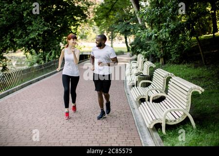 Tiro orizzontale di giovane coppia multirazziale, indossando t-shirt bianche e pantaloni neri, jogging nel vicolo del parco cittadino, al mattino presto con l'alba dentro Foto Stock