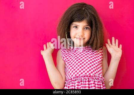 Bambina bruna con le mani in alto nell'aria, che guarda sorpreso o perso per le parole. Isolato su sfondo rosa Foto Stock