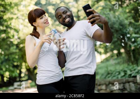 Amici multietnici moderni e gioiosi si accoppiano con bottiglie d'acqua, facendo una pausa per scattare foto selfie sullo smartphone, in piedi nel verde urbano del parco durante lo sport Foto Stock