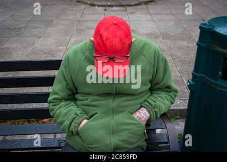 Street reportage, un uomo addormentato su una panchina nella città di Cirencester indossando un cappuccio rosso e bicchieri. Foto Stock