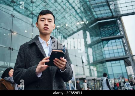 Giovane bell'uomo d'affari asiatico che guarda con la fotocamera mentre si trova in piedi con lo smartphone sulla strada vicino all'aeroporto Foto Stock