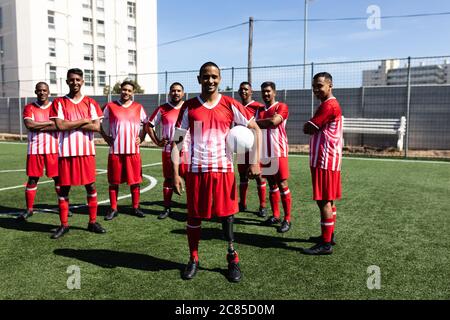 Ritratto di una squadra multietnica di cinque giocatori di calcio di lato che indossano una squadra di allenamento in un campo sportivo sotto il sole, in piedi tenendo una palla Foto Stock