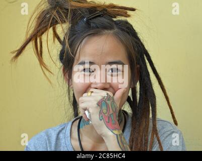 Giovane donna tailandese con lunghi e solo semi-rifiniti dreads Rastafarian posa per la macchina fotografica e copre la sua bocca con la sua mano sinistra tatuata. Foto Stock