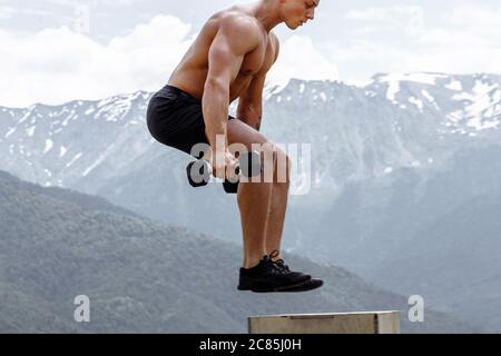 Motion shot di bel forte uomo che fa allenamento funzionale su cielo e montagna sfondo, eseguendo Box salto esercizio all'aperto. Foto Stock