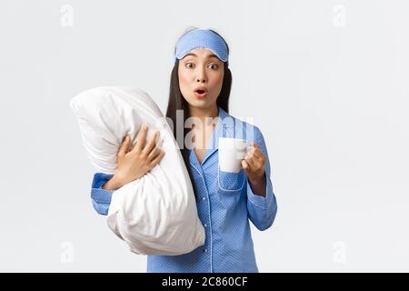 Stile di vita mattutino, colazione e concetto di gente. Sorpresa e curiosa ragazza asiatica cutr in maschera addormentata e pigiama, tenendo una tazza di caffè e cuscino Foto Stock