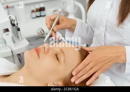 Primo piano di una bella giovane donna che si trova con gli occhi chiusi mentre beautician professionista che esegue microdermoabrasione esfoliante cosmetare pelle Foto Stock