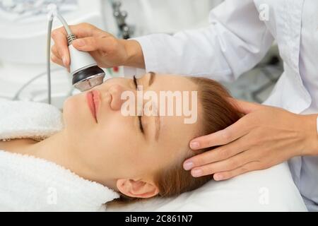 Bella giovane donna sorridente mentre riceve trattamento viso ultrasonico cura della pelle da cosmetologo professionale benessere coccole dermatologia fresco Foto Stock