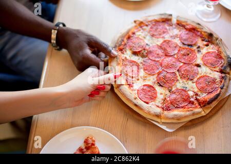 Cena estiva o pranzo al bar interno. Piatto di mani di persone che prendono fresco pepperoni pizza italiana con fette di salame su legno bianco Foto Stock