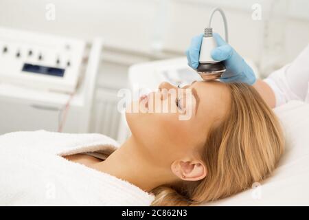 Splendida giovane donna sdraiata con gli occhi chiusi rilassandosi mentre riceve trattamento viso ultrasonico presso la moderna clinica di bellezza clinica salone terapeuta thera Foto Stock