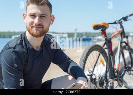 Ritratto di un atleta di ciclista che riposa dopo un giro in bicicletta usando uno smartphone in mano. All'aperto. Sport primaverili. Il ragazzo è venticinque voi Foto Stock