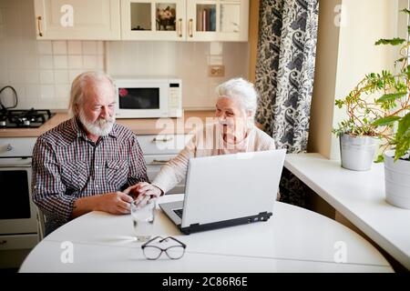 Bella coppia anziana allegra seduta al tavolo da cucina e utilizzando tablet digitale per pagare il costo di utilità. Stile di vita senior, persone mature e tec Foto Stock