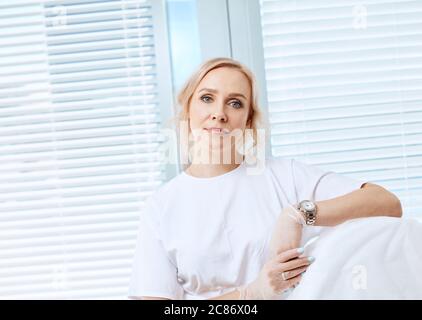 Amichevole giovane donna caucasica guardando medico in uniforme bianca guardando la macchina fotografica sopra sfondo finestra Foto Stock