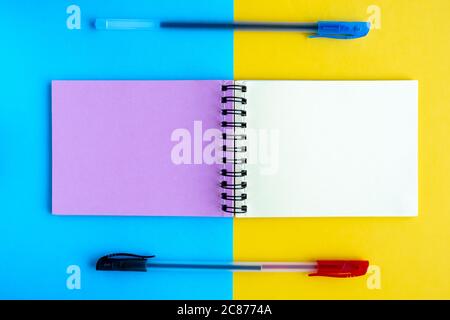 Un notebook con pagine multicolore e due penne a sfera giacciono su uno sfondo giallo e blu. Vista dall'alto, spazio per testo, disposizione piatta. Foto Stock